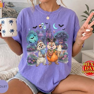 May include: A light purple short-sleeve t-shirt featuring a graphic print of two cartoon chipmunks in Halloween costumes, one dressed as a mummy and the other as a pumpkin, standing in front of a theme park castle, trees, and bats.