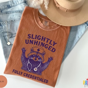 May include: An orange t-shirt with the text "SLIGHTLY UNHINGED FULLY CREDENTIALED" and a purple raccoon wearing a nurse's hat and stethoscope. The shirt is folded on top of blue jeans, with a tan hat and a gold necklace visible.