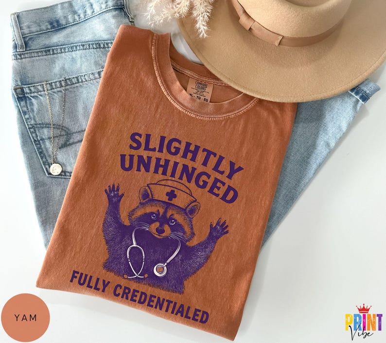 May include: An orange t-shirt with the text "SLIGHTLY UNHINGED FULLY CREDENTIALED" and a purple raccoon wearing a nurse's hat and stethoscope. The shirt is folded on top of blue jeans, with a tan hat and a gold necklace visible.
