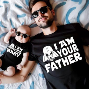 May include: A black t-shirt with the text "I AM YOUR FATHER" and a Darth Vader graphic, worn by an adult. A baby wears a matching black onesie with "I AM YOUR BABY" and a Darth Vader graphic. Both wear sunglasses.