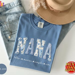 May include: A blue crewneck t-shirt featuring the word NANA in large, block-style letters with varied blue and white floral, gingham, and polka dot patterns. Below the letters, the names bella, maria, angel, and zoi are printed in white script.