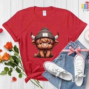 Op de afbeelding: Rood T-shirt met een cartoon Highland-koe met een muts. De koe heeft lang, ruig bruin haar, grote hoorns en een geruit overhemd. Het shirt wordt gecombineerd met een blauwe spijkerbroek en witte sneakers.