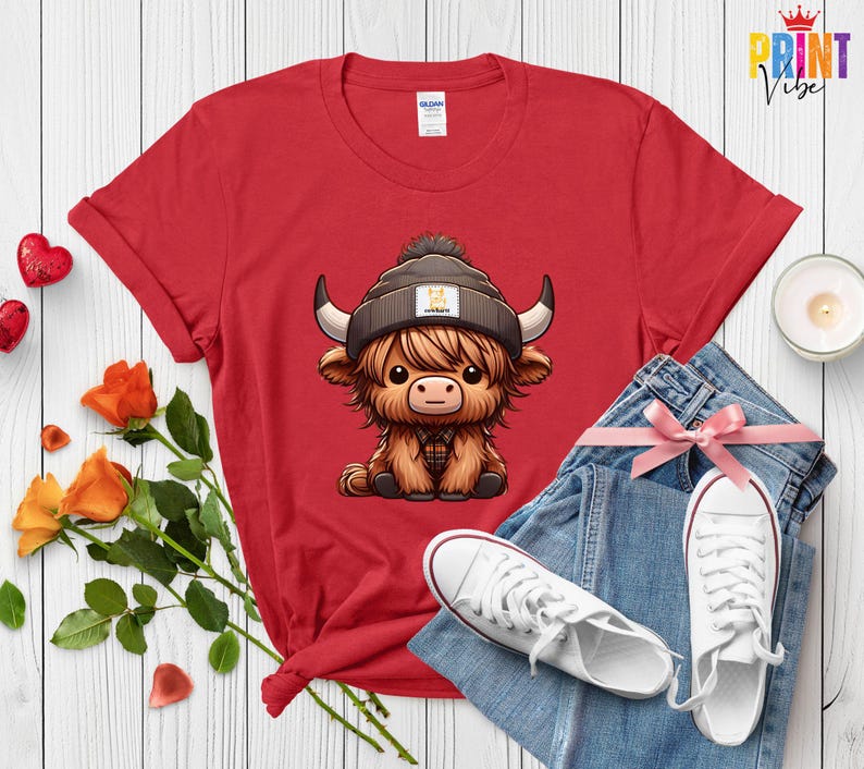Op de afbeelding: Rood T-shirt met een cartoon Highland-koe met een muts. De koe heeft lang, ruig bruin haar, grote hoorns en een geruit overhemd. Het shirt wordt gecombineerd met een blauwe spijkerbroek en witte sneakers.