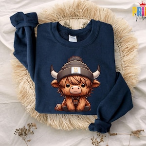 Op de afbeelding: Marineblauwe sweatshirt met een cartoon Highland koe-ontwerp. De koe draagt een muts en een geruit overhemd. De afbeelding toont de sweatshirt op een geweven, beige oppervlak. De koe heeft bruine vacht en grote hoorns.