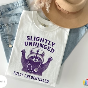 May include: White t-shirt featuring the text "SLIGHTLY UNHINGED FULLY CREDENTIALED" and a purple raccoon graphic wearing a nurse's hat and stethoscope. The shirt is shown with blue jeans, a gold necklace, and a tan hat.