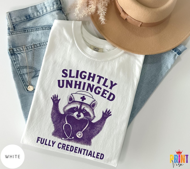 May include: White t-shirt featuring the text "SLIGHTLY UNHINGED FULLY CREDENTIALED" and a purple raccoon graphic wearing a nurse's hat and stethoscope. The shirt is shown with blue jeans, a gold necklace, and a tan hat.