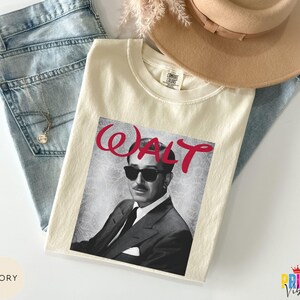 May include: Ivory-colored t-shirt featuring a black and white image of a man wearing sunglasses, with the name "WALT" in red script. The shirt is laid on top of blue jeans and a tan hat.