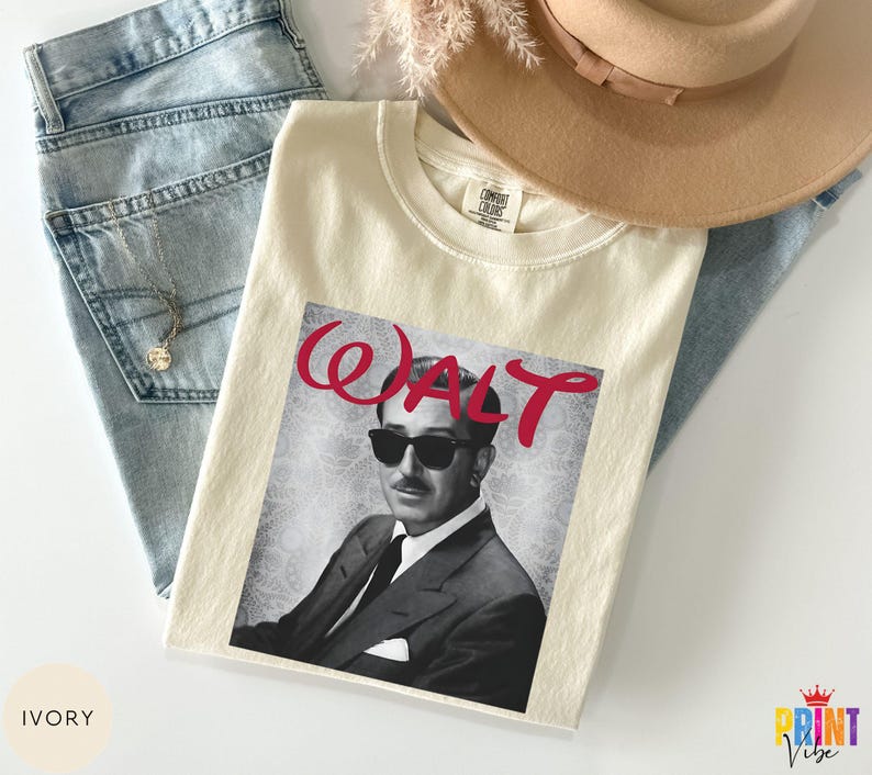 May include: Ivory-colored t-shirt featuring a black and white image of a man wearing sunglasses, with the name "WALT" in red script. The shirt is laid on top of blue jeans and a tan hat.