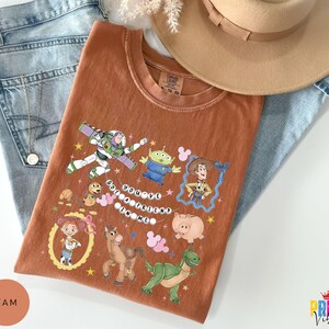May include: A rust-colored short-sleeve t-shirt featuring a graphic print of various animated characters including a space ranger, a cowboy, a cowgirl, a green alien, a dinosaur, a pig, a horse, and a dog, with the text "YOU'VE GOT A FRIEND IN ME".
