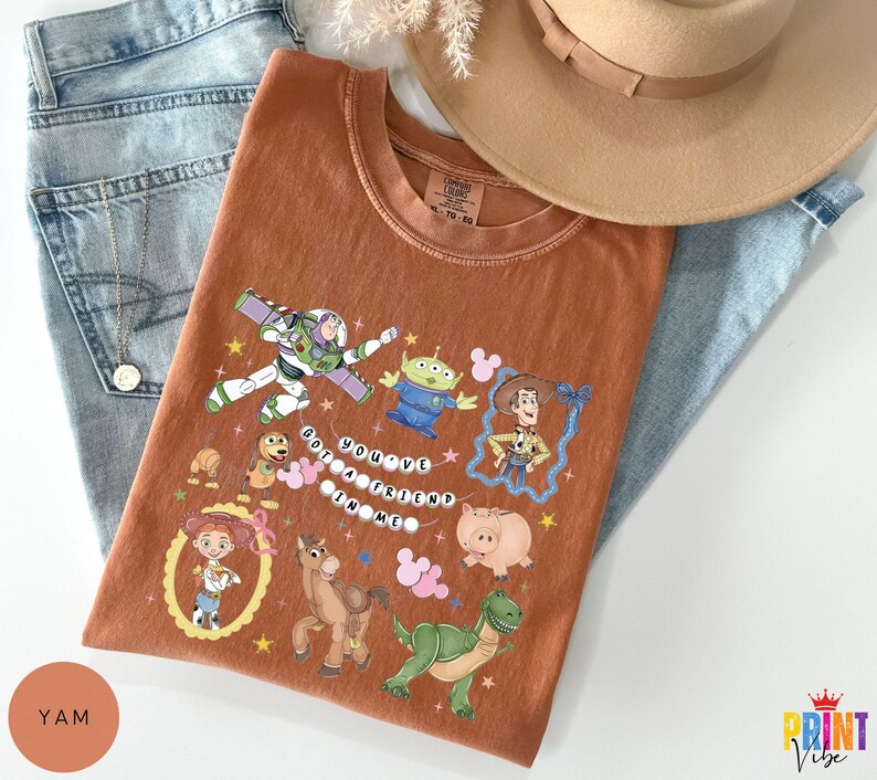 May include: A rust-colored short-sleeve t-shirt featuring a graphic print of various animated characters including a space ranger, a cowboy, a cowgirl, a green alien, a dinosaur, a pig, a horse, and a dog, with the text "YOU'VE GOT A FRIEND IN ME".