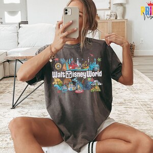 Puede incluir: Camiseta gris oscuro con un gr&aacute;fico colorido de los lugares emblem&aacute;ticos de Walt Disney World. El texto "Walt Disney World" se muestra de forma destacada en la parte delantera. La camiseta tiene mangas cortas y un corte holgado.