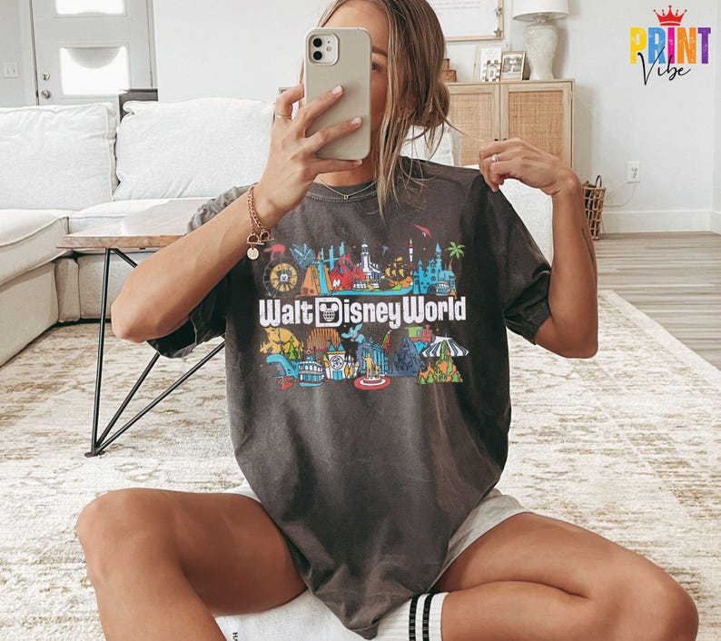 Puede incluir: Camiseta gris oscuro con un gr&aacute;fico colorido de los lugares emblem&aacute;ticos de Walt Disney World. El texto "Walt Disney World" se muestra de forma destacada en la parte delantera. La camiseta tiene mangas cortas y un corte holgado.