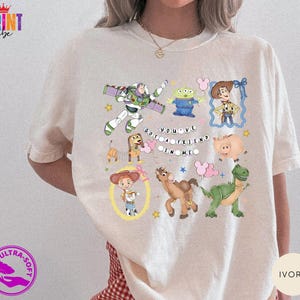 May include: An ivory t-shirt featuring a colorful graphic print of various animated characters including a space ranger, a green alien, a cowboy, a dachshund, a pig, a cowgirl, a horse, and a dinosaur, with the text "YOU'VE GOT A FRIEND IN ME".
