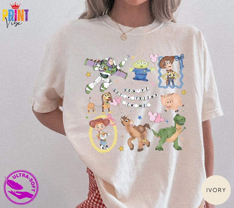 May include: An ivory t-shirt featuring a colorful graphic print of various animated characters including a space ranger, a green alien, a cowboy, a dachshund, a pig, a cowgirl, a horse, and a dinosaur, with the text "YOU'VE GOT A FRIEND IN ME".