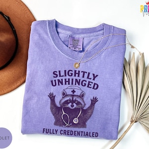 May include: A violet Comfort Colors t-shirt featuring a raccoon graphic wearing a nurse's hat and stethoscope. The text reads "Slightly Unhinged Fully Credentialed". A gold necklace and a brown hat are also in the image.