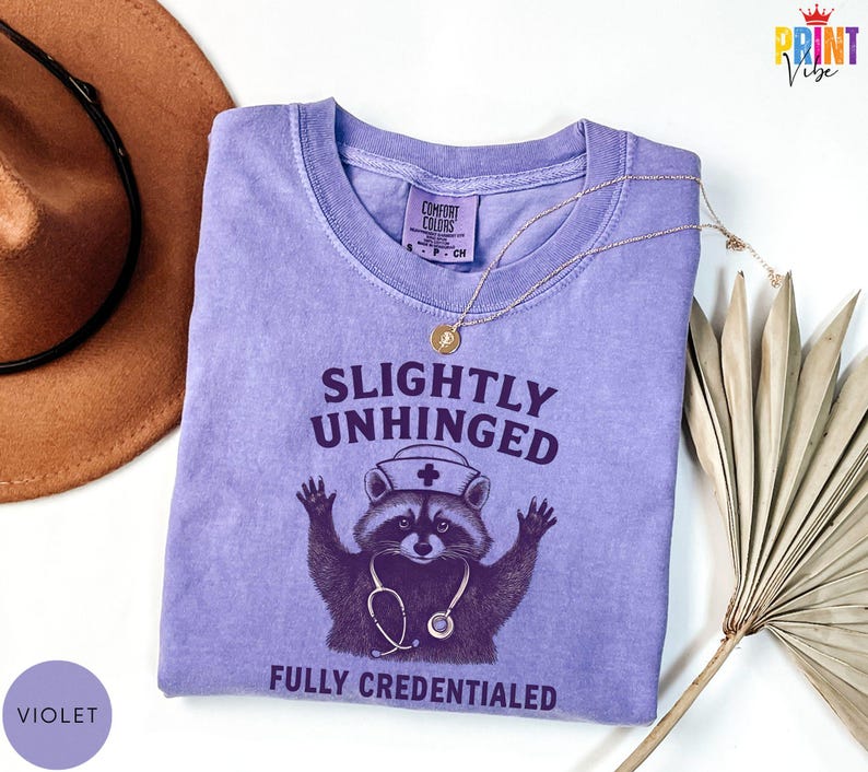 May include: A violet Comfort Colors t-shirt featuring a raccoon graphic wearing a nurse's hat and stethoscope. The text reads "Slightly Unhinged Fully Credentialed". A gold necklace and a brown hat are also in the image.