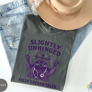 May include: A grey t-shirt featuring the text "SLIGHTLY UNHINGED FULLY CREDENTIALED" and a purple raccoon wearing a nurse's hat and stethoscope. The shirt is on blue jeans and a tan hat.
