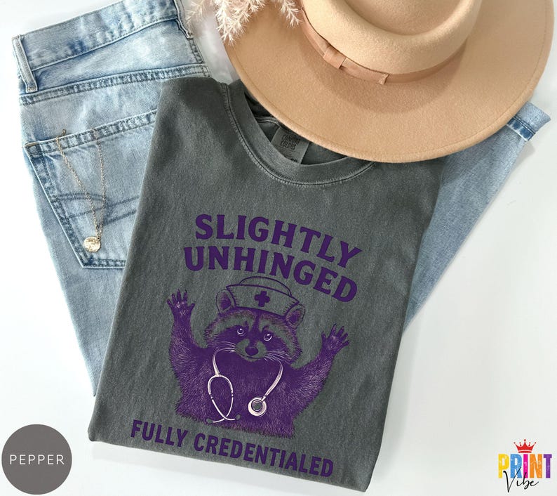 May include: A grey t-shirt featuring the text "SLIGHTLY UNHINGED FULLY CREDENTIALED" and a purple raccoon wearing a nurse's hat and stethoscope. The shirt is on blue jeans and a tan hat.