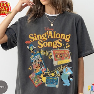May include: A charcoal gray t-shirt featuring a graphic print with the text Disney Sing Along Songs. The design includes illustrations of Jiminy Cricket, a Disneyland Fun cassette tape, a VHS tape, and a blue bird character with musical notes.