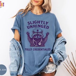 May include: A light blue t-shirt featuring the text "SLIGHTLY UNHINGED FULLY CREDENTIALED" and a purple raccoon wearing a nurse's hat and stethoscope. The shirt is a light blue colour.