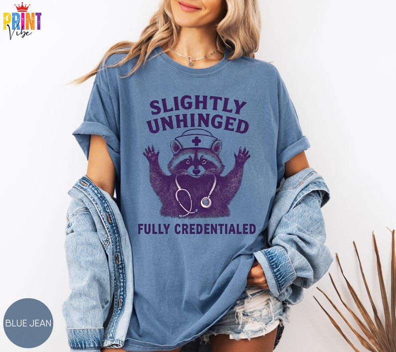 May include: A light blue t-shirt featuring the text "SLIGHTLY UNHINGED FULLY CREDENTIALED" and a purple raccoon wearing a nurse's hat and stethoscope. The shirt is a light blue colour.