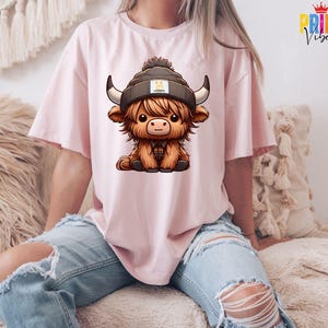 Op de afbeelding: Lichtroze T-shirt met een cartoon illustratie van een Schotse Hooglander met een muts. De koe heeft grote hoorns en een bruine vacht. De persoon draagt een gescheurde blauwe jeans.