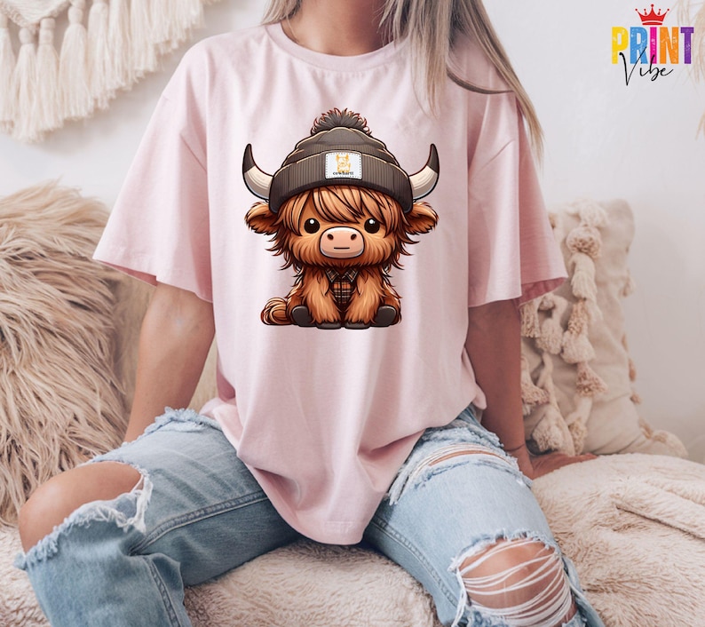 Op de afbeelding: Lichtroze T-shirt met een cartoon illustratie van een Schotse Hooglander met een muts. De koe heeft grote hoorns en een bruine vacht. De persoon draagt een gescheurde blauwe jeans.