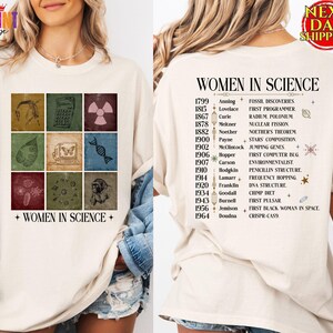 May include: A cream-colored t-shirt featuring a front graphic with nine squares displaying scientific icons and the text "WOMEN IN SCIENCE." The back displays a list of names, years, and scientific achievements under the heading "WOMEN IN SCIENCE."