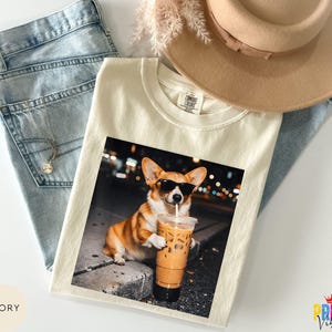 May include: Ivory-colored t-shirt featuring a graphic of a corgi wearing sunglasses and holding an iced coffee. The shirt is laid on top of blue jeans, a necklace, and a tan hat. The shirt has a Comfort Colors tag.