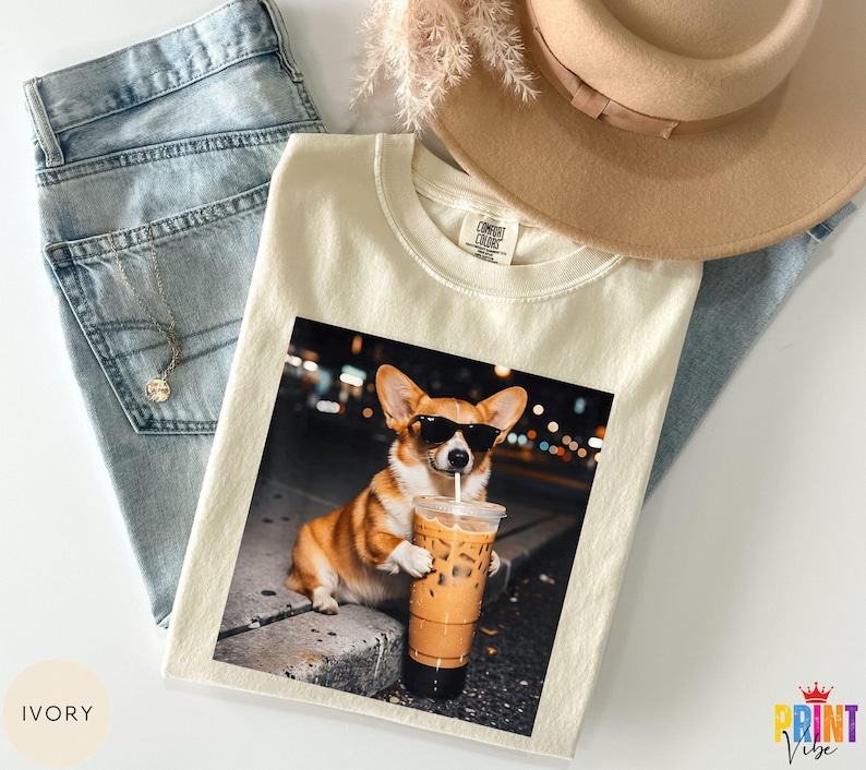 May include: Ivory-colored t-shirt featuring a graphic of a corgi wearing sunglasses and holding an iced coffee. The shirt is laid on top of blue jeans, a necklace, and a tan hat. The shirt has a Comfort Colors tag.