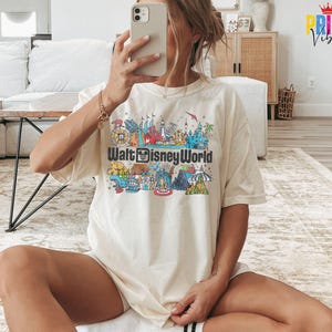 Puede incluir: Camiseta color crema con un gr&aacute;fico colorido de los monumentos de Walt Disney World y el logotipo del parque. El dise&ntilde;o incluye ilustraciones de varias atracciones y edificios, creando una escena vibrante y detallada.