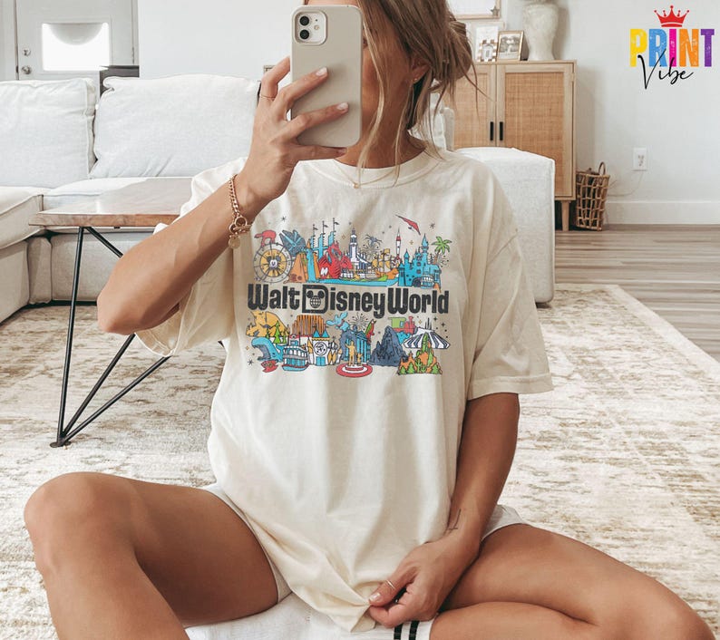 Puede incluir: Camiseta color crema con un gr&aacute;fico colorido de los monumentos de Walt Disney World y el logotipo del parque. El dise&ntilde;o incluye ilustraciones de varias atracciones y edificios, creando una escena vibrante y detallada.