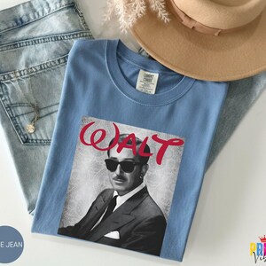 May include: A light blue t-shirt featuring a black and white image of a man wearing sunglasses and a suit, with the name "WALT" in red script. The shirt is folded on top of blue jeans.