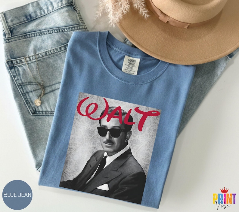 May include: A light blue t-shirt featuring a black and white image of a man wearing sunglasses and a suit, with the name "WALT" in red script. The shirt is folded on top of blue jeans.