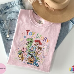 May include: A light pink t-shirt featuring a colorful graphic print of Toy Story characters including Woody, Buzz Lightyear, Jessie, Rex, Slinky Dog, Mr. Potato Head, and the Aliens, with the text Toy Story and scribbled star and circle designs.
