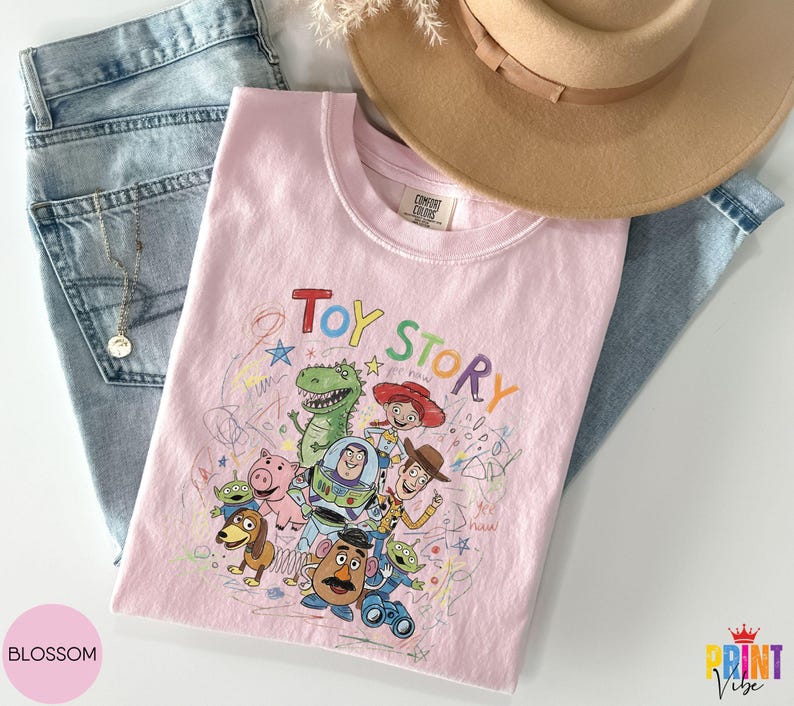 May include: A light pink t-shirt featuring a colorful graphic print of Toy Story characters including Woody, Buzz Lightyear, Jessie, Rex, Slinky Dog, Mr. Potato Head, and the Aliens, with the text Toy Story and scribbled star and circle designs.