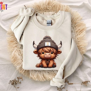 Op de afbeelding: Cr&egrave;mekleurige sweatshirt met een cartoon Highland-koe met een grijze beanie met een geel vierkant logo. De koe heeft bruine vacht, zwarte hoorns en een geruit shirt. De sweatshirt ligt op een geweven mat.