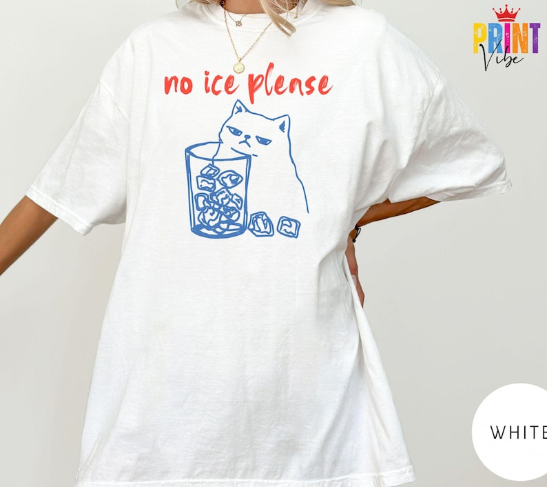 May include: White t-shirt with a blue illustration of a grumpy cat next to a glass of ice cubes. The text "no ice please" is printed in red above the cat and glass. The shirt has short sleeves.