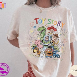 May include: An ivory short-sleeve t-shirt featuring a colorful graphic print of Toy Story characters including Woody, Buzz Lightyear, Jessie, Rex, Hamm, Slinky Dog, Mr. Potato Head, and Aliens, surrounded by multi-colored scribbles and text.