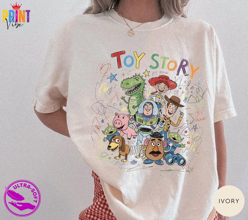 May include: An ivory short-sleeve t-shirt featuring a colorful graphic print of Toy Story characters including Woody, Buzz Lightyear, Jessie, Rex, Hamm, Slinky Dog, Mr. Potato Head, and Aliens, surrounded by multi-colored scribbles and text.