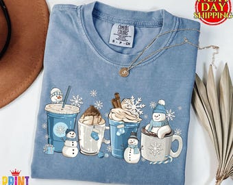 May include: A blue short-sleeved t-shirt featuring a graphic print of four winter-themed beverage containers, including cups and mugs, decorated with snowmen, snowflakes, and whipped cream. The shirt has a Comfort Colors brand label at the neck.