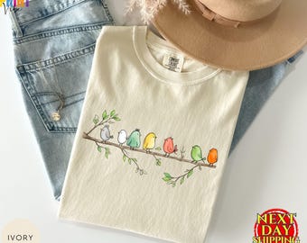 Może przedstawiać: T-shirt w kolorze kości słoniowej z grafiką przedstawiającą siedem kolorowych ptaków siedzących na brązowej gałęzi drzewa z zielonymi liśćmi. Koszulka leży na jasnoniebieskich dżinsach i jest uzupełniona beżowym kapeluszem oraz srebrnym naszyjnikiem.