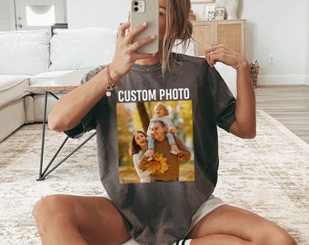 May include: A charcoal gray short-sleeve t-shirt featuring the text CUSTOM PHOTO printed in white above a square graphic of a family portrait. The graphic shows a person carrying a child on their shoulders with another person standing beside them.