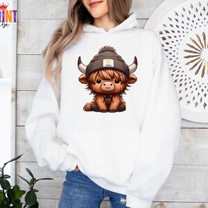Op de afbeelding: Witte hoodie met een cartoon illustratie van een Schotse Hooglander koe met een muts. De koe heeft bruine vacht, een geruit overhemd en een grijze muts met een kleine patch. De hoodie heeft een kangoeroezak.
