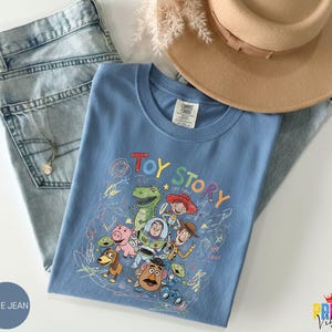 May include: A blue t-shirt featuring a colorful graphic print of Toy Story characters including Woody, Buzz Lightyear, Rex, and Jessie, surrounded by scribbled crayon-style drawings. The shirt is paired with light blue denim jeans and a tan hat.