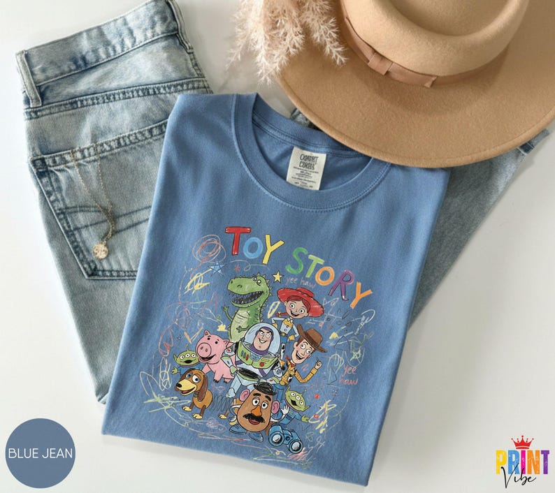 May include: A blue t-shirt featuring a colorful graphic print of Toy Story characters including Woody, Buzz Lightyear, Rex, and Jessie, surrounded by scribbled crayon-style drawings. The shirt is paired with light blue denim jeans and a tan hat.