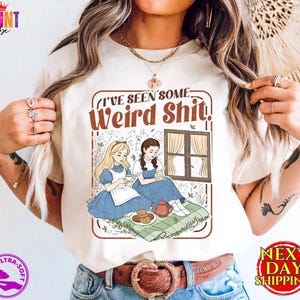 May include: Beige t-shirt with a graphic that reads "I've Seen Some Weird Shit." The graphic features two cartoon women in blue dresses, seated near a window. The shirt has a round neck and short sleeves. The words "Ultra-Soft" are printed on the shirt.