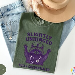 May include: An olive green t-shirt with the text "SLIGHTLY UNHINGED FULLY CREDENTIALED" and a purple raccoon graphic wearing a nurse's hat and stethoscope. The shirt is laid on top of blue jeans and a tan hat.