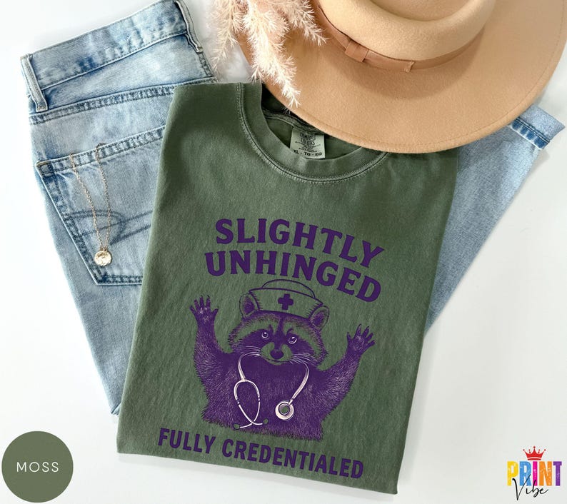 May include: An olive green t-shirt with the text "SLIGHTLY UNHINGED FULLY CREDENTIALED" and a purple raccoon graphic wearing a nurse's hat and stethoscope. The shirt is laid on top of blue jeans and a tan hat.