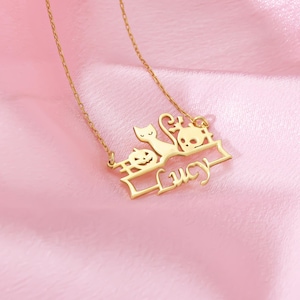 May include: A gold-colored necklace with a Halloween theme. The pendant features a cat, a skull, and a jack-o'-lantern, with the name "Lucy" in cursive script. The necklace is displayed on a pink satin surface.